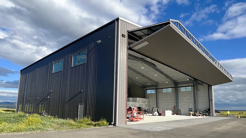 Exterior lefthand view of Schweiss bifold hangar door installed on gray tin hangar shown open Exterior lefthand view of Schweiss bifold hangar door installed on gray tin hangar shown open