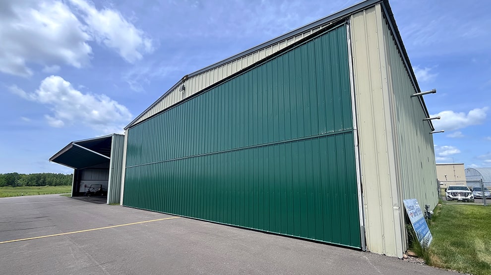 Green Schweiss bifold lift strap hangar doors installed on two olive colored hangars Green Schweiss bifold lift strap hangar doors installed on two olive colored hangars
