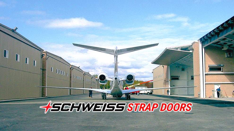 Schweiss strap doors logo shown underneath rear end of jet plane with hangars surrounding the left and right of the camera Schweiss strap doors logo shown underneath rear end of jet plane with hangars surrounding the left and right of the camera