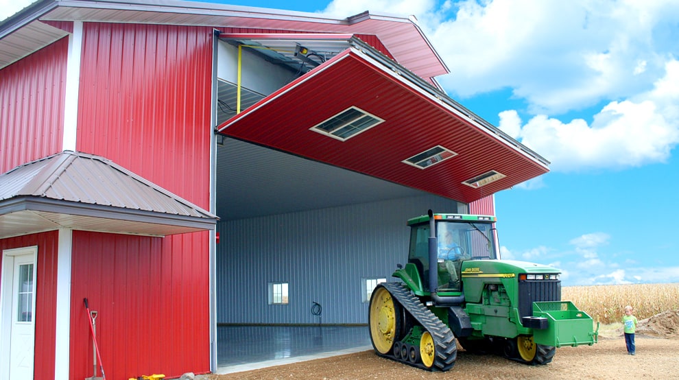 Green tractor parked outside of barn-like ag building fitted with a Schweiss liftstrap bifold door shown open Green tractor parked outside of barn-like ag building fitted with a Schweiss liftstrap bifold door shown open